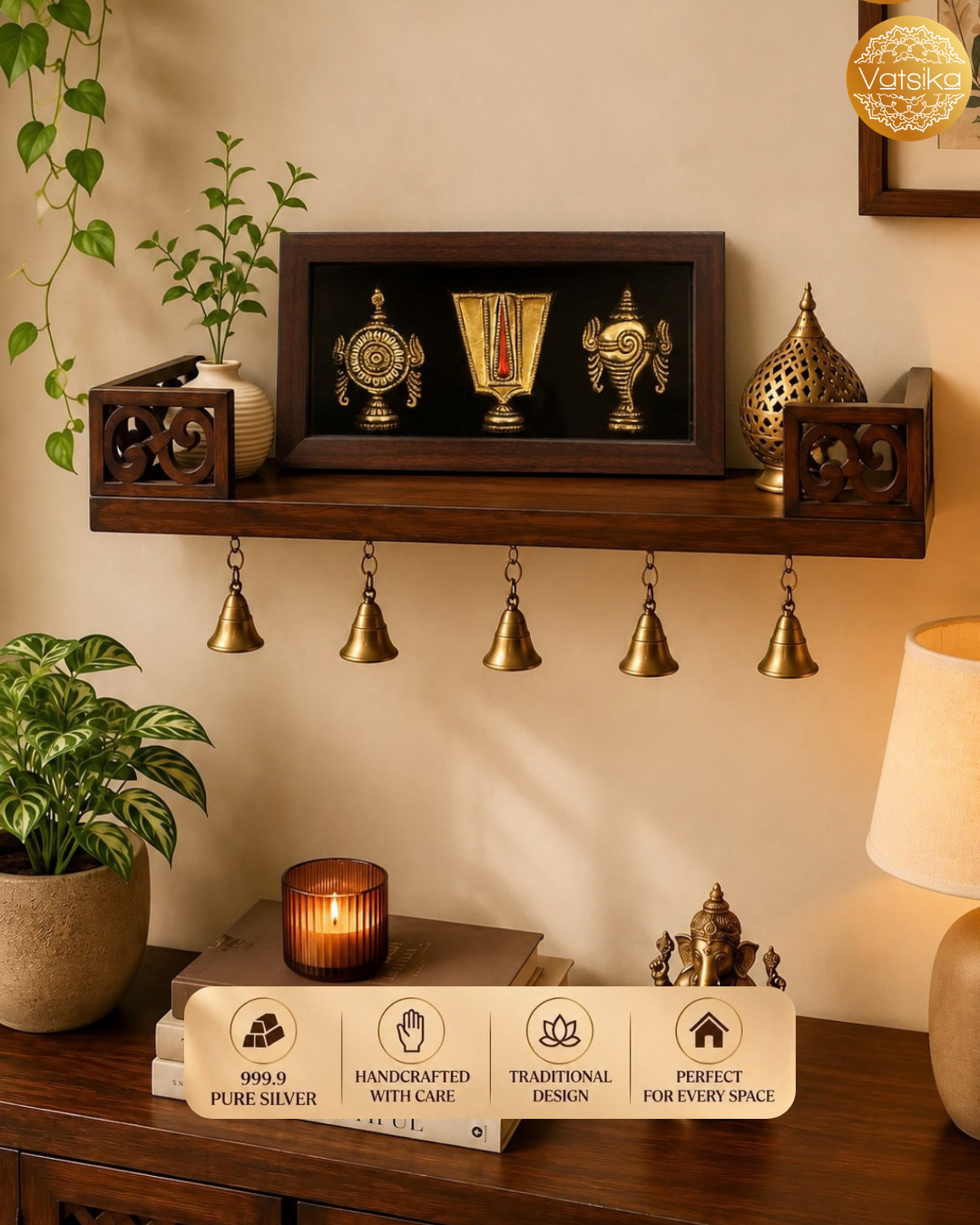 A decorative Silver Shanku Chakra Nama wall-mounted shelf in a warm indoor setting, a carved side box, and a perforated decorative object above five hanging brass bells. A lit candle, books, a small figurine, and a vase with greenery sit below on a wooden surface, with a lamp on the right and a Vatsika brand badge in the top-right corner.